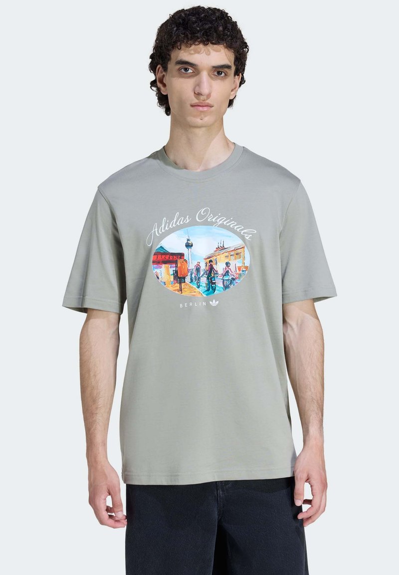 Ljust grå kortärmad t-shirt med en färgglad grafisk tryck i oval form, texten "Adidas Originals" och "Berlin" nedanför.