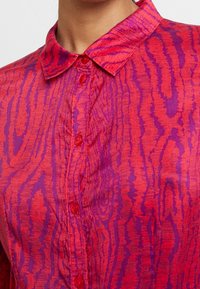 Blusa rossa e viola con motivo, colletto e chiusura con bottoni. Il tessuto presenta un design testurizzato e ondulato con una leggera lucentezza.