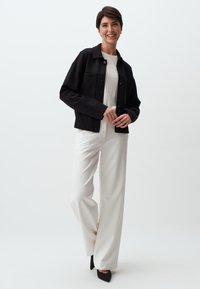 Zwarte cropped jas met drukknoopsluiting, gecombineerd met een witte top en vloeiende witte broek. Het model draagt zwarte puntige hakken. Eenvoudig, strak design.