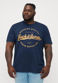 Marineblaues Kurzarm-T-Shirt aus Baumwolle. Mit einem großen, orangen Grafikdruck mit dem Text "Jack & Jones" und "Denim Supply Co."