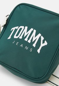 Zelená látková taška přes rameno se hladkým povrchem, na které je bílý nápis "TOMMY" a "JEANS". Obsahuje pruhovaný nastavitelný popruh.