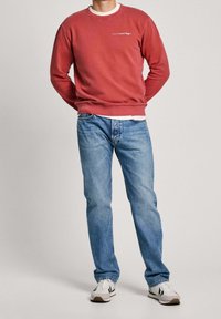 Rotes Sweatshirt mit strukturierter Stoffoberfläche und gerippten Bündchen, darüber ein weißes Hemd getragen. Kombiniert mit blauen Jeans und weißen Turnschuhen.