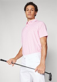 Short-sleeve golf polo shirt in pink with a geometric pattern, paired with white pants. The model holds a golf club, showcasing athletic fit.