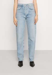 Henkilö pukeutuneena vaaleansiniset korkeavyötäröiset suoralahkeiset farkut, joissa hienovarainen kulutusefekti, ja mustat kärjestä terävät nilkkurit seisoo yksiväristä taustaa vasten.