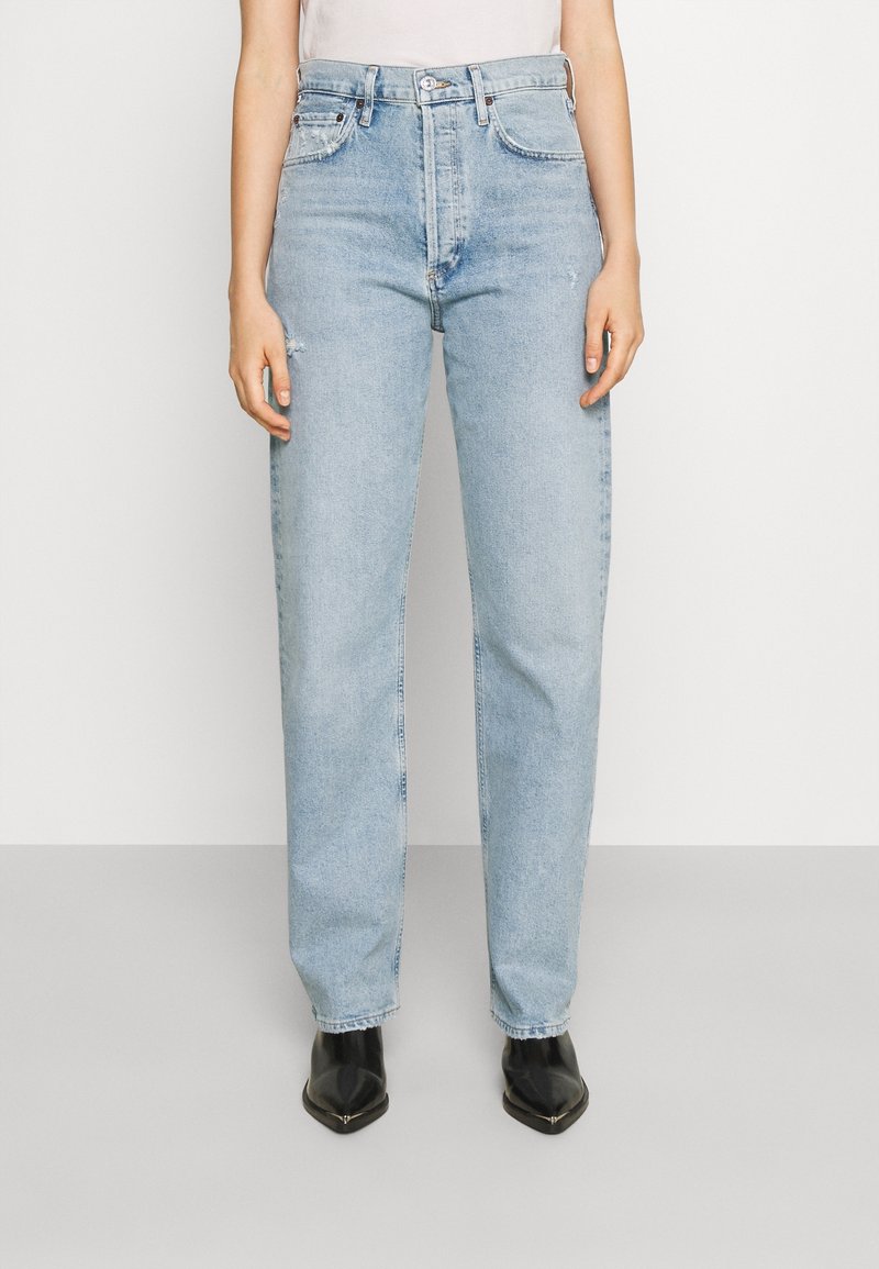 Henkilö pukeutuneena vaaleansiniset korkeavyötäröiset suoralahkeiset farkut, joissa hienovarainen kulutusefekti, ja mustat kärjestä terävät nilkkurit seisoo yksiväristä taustaa vasten.