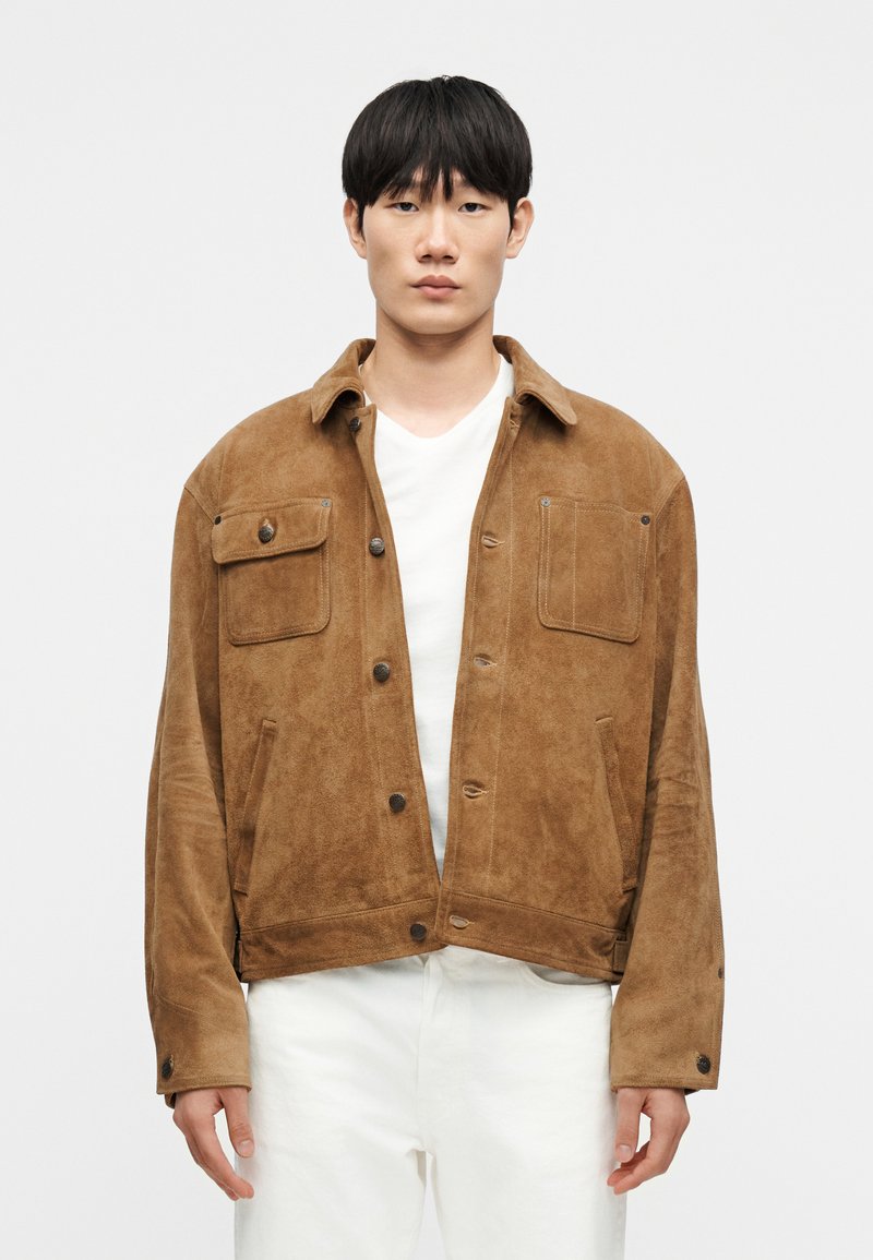 Καφέ σατέν σακάκι με γιακά, δύο μπρ οστινές τσέπες, κλείσιμο με κουμπί πατιλέτα και άνετη εφαρμογή, φορεμένο πάνω από λευκή μπλούζα.