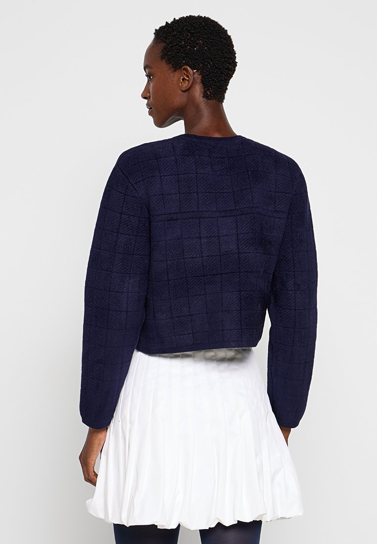 Granatowy krótki sweter wykonany z teksturowanej tkaniny w kratkę, z długimi rękawami i okrągłym dekoltem, noszony na białej plisowanej spódnicy.