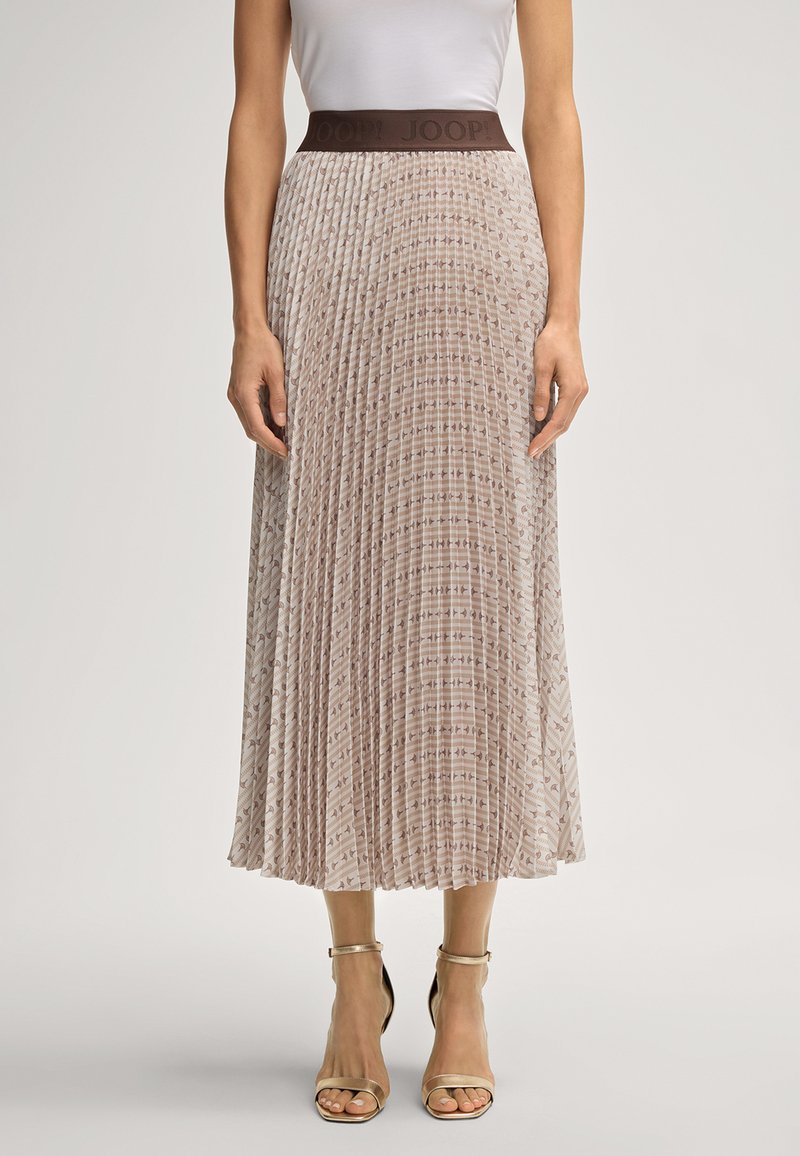 Mulher vestindo uma saia midi plissada bege com um design estampado e uma faixa escura na cintura, combinada com uma blusa branca e sapatos de salto com tiras no tornozelo bege.