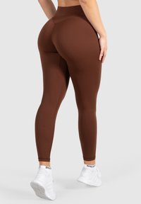 Bruine high-waisted leggings van rekbaar materiaal, met een gladde textuur en een getailleerd ontwerp. Witte sportschoenen met drie zichtbare strepen.