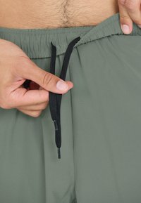 Manliga händer justerar svart snöre på olivgröna shorts med elastisk midja, vilket visar en del av torse med kroppshår.