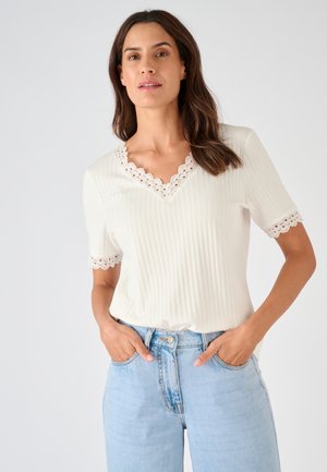 Femme aux longs cheveux bruns portant un haut côtelé à manches courtes blanc avec bordure en dentelle et un jean bleu clair, les mains dans les poches, devant un fond uni.