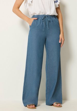 Kvinde iført blå denimbukser med vidde og snøre i taljen samt hvide sandaler, kombineret med en hvid bluse med blå broderi.