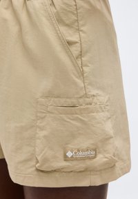 Μπεζ σορτς με πλαϊνή τσέπη και το έμβλημα της Columbia Sportswear Company στην τσέπη, φορεμένα σε σκουρόχρωμο πόδι.