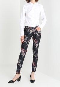 Pantalons noirs à motif floral avec des nuances de bourgogne et de gris ; design ajusté avec poches, associés à une chemise blanche à boutons et des talons noirs.