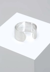 Sølvmetal cuff-ring med en glat, mat finish og åben design med buede kanter, vist på en hævet hvid platform.