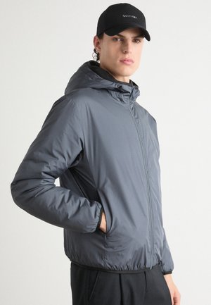 Graue, mit Reißverschluss versehene Jacke mit Kapuze. Hergestellt aus glattem, leichtem Stoff. Verfügt über Seitentaschen und elastische Bündchen. Getragen über schwarzen Hosen.