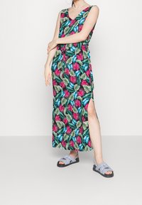 Kvinna klädd i en ärmlös, färgglad maxi-klänning med tropiskt mönster och sidoklipp, bärande ljusblå knutna sandaletter, stående mot en vit vägg.