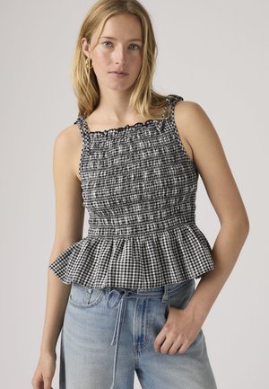 Frau trägt schwarzen und weißen ärmellosen Oberteil mit Rüschenabschluss und hellblaue High-Waist-Jeans vor einfachem Hintergrund.