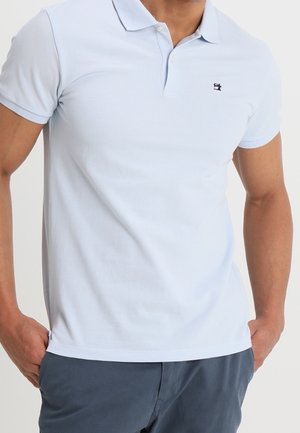 Homme portant un polo bleu clair à manches courtes avec un petit logo foncé sur la poitrine et un pantalon bleu foncé, debout les mains dans les poches.