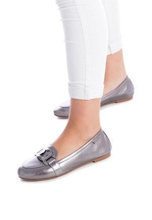 Fødder iført metallisk-sølv flade loafers med spænde, kombineret med hvide opkørte skinny bukser på en hvid baggrund.