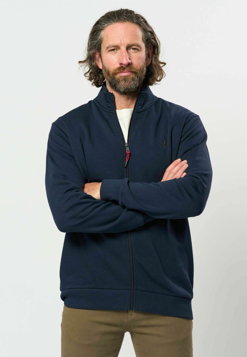 Homme avec barbe et cheveux mi-longs, portant une veste zippée bleu marine et un pantalon marron, debout les bras croisés devant un fond uni.