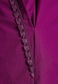 Gros plan sur un tissu violet profond avec une bordure tressée décorative présentant de petits éventails texturés le long d’une couture en zigzag.