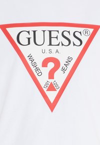 White t-shirt featuring a large red triangle logo with "GUESS" and "WASHED JEANS" text, accompanied by a question mark and numbers 1201 and 1203.