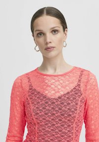 Top in pizzo di un vivace rosa neon con maniche lunghe e colletto rotondo. Motivo testurizzato con uno strato sottostante scuro in contrasto visibile.