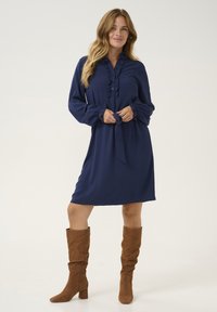 Robe bleu marine avec un col froncé, des détails en boutons et de longues manches bouffantes. Associée à des bottes en suède marron hauteur genoux.