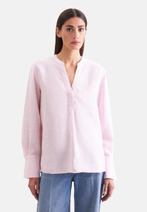 Vrouw met stijl donker haar, die een lichtroze blouse met lange mouwen en V-hals en blauwe spijkerbroek draagt, staand tegen een effen achtergrond.