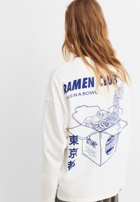 Bijela košulja dugih rukava s plavim grafičkim printom zdjele ramena i tekstom "RAMEN KLUB #POTPARA U ZDJELI" na poleđini.