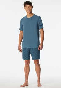 Helleblaue Kurzarmbluse aus glattem Stoff, kombiniert mit gemusterten dunkelblauen Shorts mit geometrischem Design.