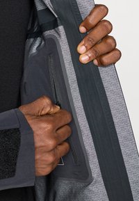 Ruky s tmavou pokožkou zipsujú vnútornú kapsu šedej a čiernej bundy s vzorovanou podšívkou a vodotesnou lepiacou páskou.