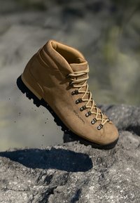 Tan Wanderschuh mit beigen Schnürsenkeln und schwarzer, robuster Sohle schwebt über felsigem Gelände im Freien.