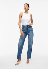 Haut sans manches blanc, jeans taille haute bleu clair, et sandales à talons beiges. Le mannequin prend la pose devant un fond blanc uni.