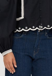 Black blouse with white scalloped lace trim and floral embroidery. Paired with dark denim jeans having topped stitching and side pockets.