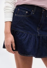 Κοντινό πλάνο ενός ατόμου που φοράει σκούρο μπλε τζιν φούστα με βολάν και μακρυμάνικο λευκό πουκάμισο με κόκκινες ρίγες, με το χέρι να ακουμπάει δίπλα του.