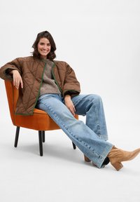 Smeđa postavljena jakna s zelenim obrubom, bež pleteni pulover, svijetloplave flare traperice i bež školske čizme, sjedi u narančastoj stolici.