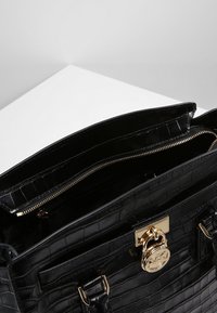 Sort håndtaske med krokodillemønster og blank finish, guldfarvet hardware, dobbelt håndtag og en lynlåslomme indeni. Åbningen er synlig.