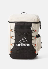 Nahrbtnik z teksturiranim bež vrhom in črnim dnom. Ima prilagodljive elastične vrvice, stranski žep in belo Adidasovo logo na sprednji strani.