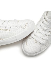 White canvas sneakers featuring gold text pattern, round toe, flat laces, and white rubber sole. Textured surface with subtle detailing.