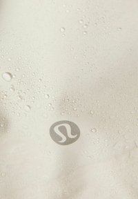 Vattentåligt tyg i ljusbeige med synliga vattendroppar. Har en silverlogotyp i nedre högra hörnet. Slät textur.