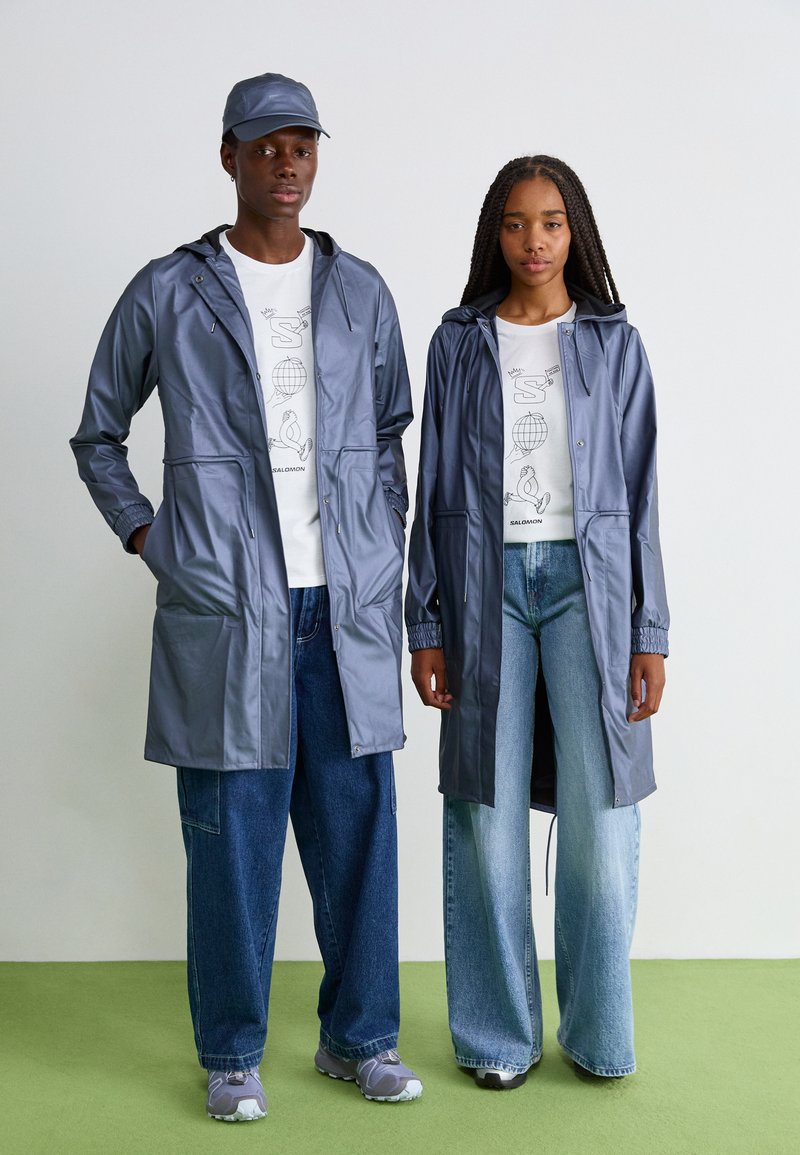 Twee modellen dragen lichtgrijze waterdichte regenjassen met trekkoorden in de capuchons, gecombineerd met graphic t-shirts en wijde jeans op een groene vloer.