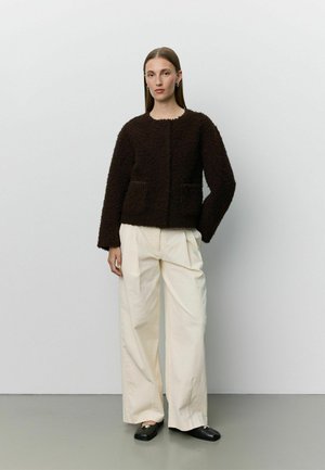 Femme portant une veste texturée marron foncé, un pantalon large crème et des chaussures noires, debout sur un fond uni gris clair.