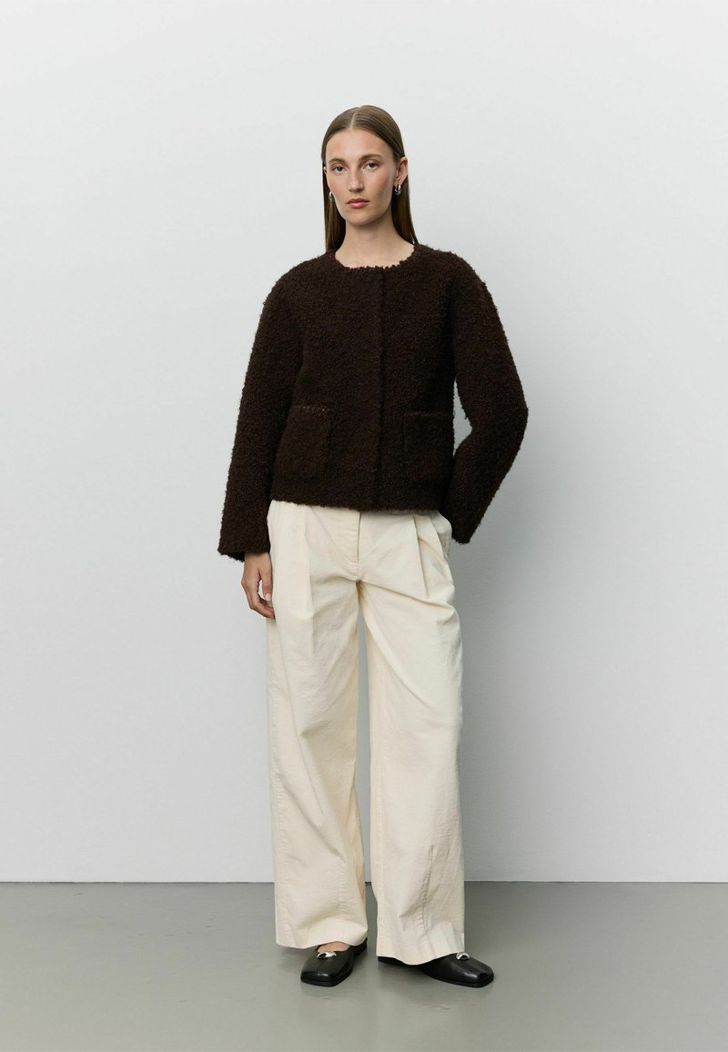 Femme portant une veste texturée marron foncé, un pantalon large crème et des chaussures noires, debout sur un fond uni gris clair.
