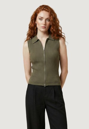 Donna con lunghi capelli rossi ricci che indossa un top senza maniche verde oliva con cerniera frontale a coste e pantaloni neri a vita alta, in piedi davanti a uno sfondo semplice.