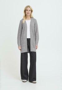 Cárdigan gris de punto con abertura delantera, mangas largas y tejido texturizado. Combinado con pantalones anchos negros y una blusa blanca, de pie sobre un fondo neutro.
