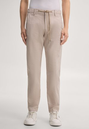 Personne portant un pantalon beige droit décontracté avec taille à cordon de serrage et des baskets blanches, debout devant un fond clair uni.