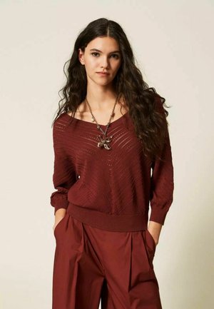 Giovane donna con capelli lunghi e ricci che indossa un maglione rosso scuro testurizzato e pantaloni larghi abbinati, accessoriata con una lunga collana con pendente.