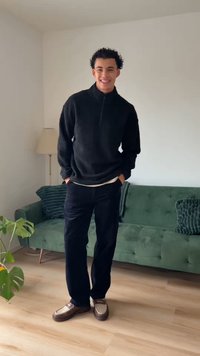 Schwarzer Fleecepullover mit Reißverschluss am Kragen, schwarze Hose und braune Schuhe mit beigefarbenen Akzenten. Grünes Sofa und Pflanze im Hintergrund.